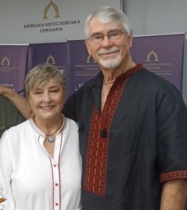 Chris and Rahnella Adsit smiling together in front of Kyiv Theological Seminary banners.