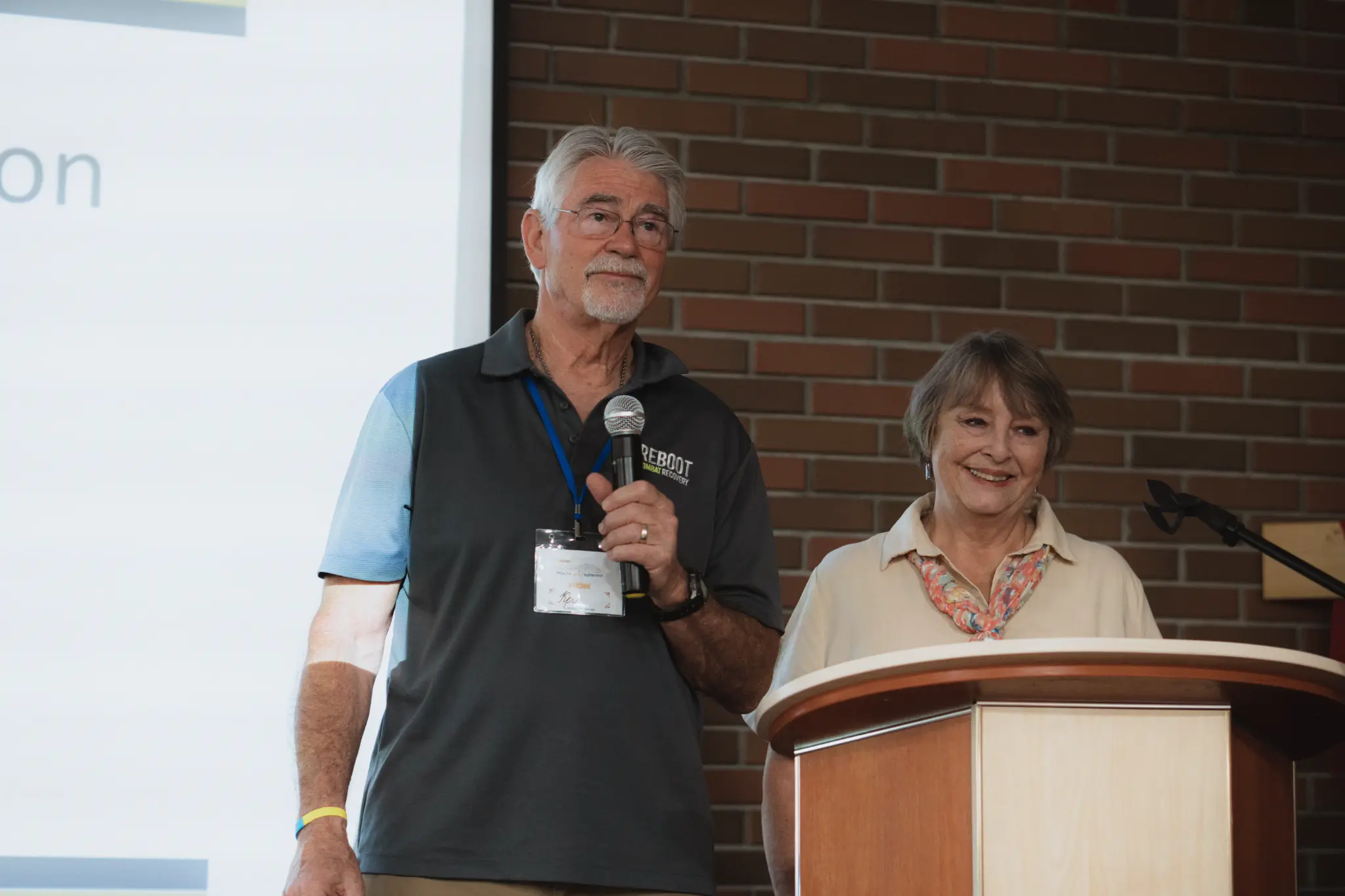 Chris speaking at a microphone while Rahnella stands at a lectern during a Reboot training.